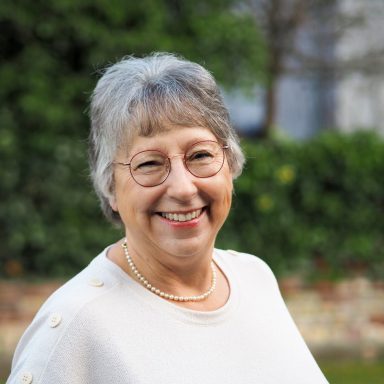 Lächelnde ältere Frau mit grauen Haaren, Brille und Perlenkette in einer grünen Umgebung.