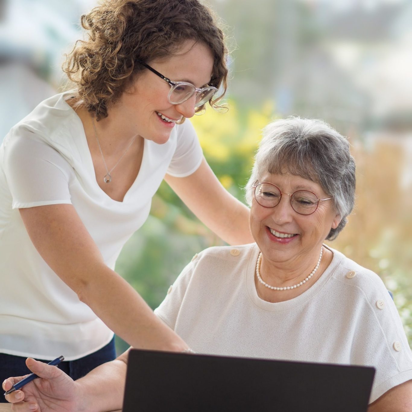 Junge Frau hilft älterer Dame beim Arbeiten am Laptop in heller Umgebung.